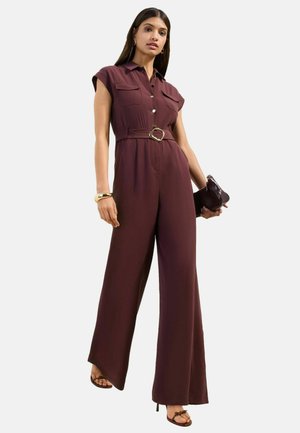 Woman wearing a sleeveless burgundy jumpsuit with wide legs, a belt, holding a matching clutch, accessorized with bracelet and hoop earrings.