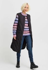 Svart vadderad lång väst med tryckknappar, ihopkopplad med en flerfärgad randig tröja i pastellrosa, lila och blått, samt blå jeans.