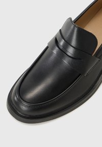 Mocassino in pelle nera con una finitura liscia e dettagli sottili sulla parte superiore, appoggiato su uno sfondo neutro.