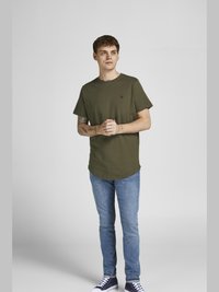 Olivgrünes T-Shirt mit kurzen Ärmeln und rundem Halsausschnitt, kombiniert mit schmal geschnittenen hellblauen Jeans und dunkelblauen Sneakers. Dezentes Logo-Detail auf der Brust.