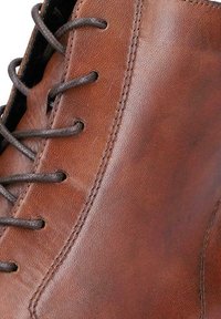Chaussure en cuir marron avec une texture lisse, bout rond et lacets sombres passés à travers des œillets ronds. Détails de couture visibles.