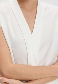 Bílá halenka s krátkým rukávem a hlubokým výstřihem ve tvaru V, hladká textura a jemný lesk, která předvádí minimalistickou estetikou.