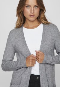 Gilet gris en tissu tricoté doux, design à ouverture devant, poignets côtelés et deux poches avant ; porté sur un haut blanc et un pantalon noir.