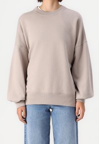 Osoba oblečená v volném béžovém svetru s balónovými rukávy a světle modrých džínách, stojící před jednoduchým bílým pozadím.