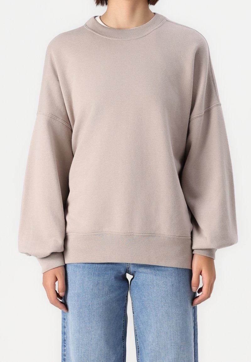 Osoba oblečená v volném béžovém svetru s balónovými rukávy a světle modrých džínách, stojící před jednoduchým bílým pozadím.