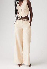 Nainen, joka on pukeutunut kremvärisiin korduroy-leveisiin housuihin ja vastaavaan lyhytlahkeiseen liiviin, mustilla remmikengillä yksinkertaista taustaa vasten.