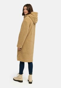 Manteau long beige en peluche avec capuche, texture douce. Porté sur un jean skinny bleu foncé et des bottes beige clair à lacets.