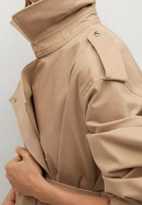 Manteau beige avec col montant, épaulettes et boutons-pression, porté par une personne ajustant la ceinture.