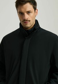 Czarna kurtka z wysokim kołnierzem, gładką teksturą, z przodem na zamek błyskawiczny i stonowanym designem. Model ma krótkie włosy i kolczyk.