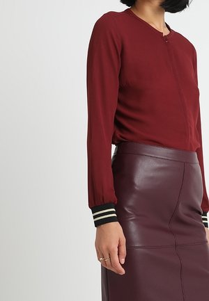 Vrouw draagt een bordeauxrode top met lange mouwen en gestreepte manchetten, en een hoogtailored donkerpaarse leren rok, staand tegen een effen achtergrond.