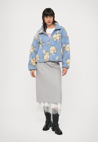 Giacca in pile blu con stampa floreale, chiusura con cerniera e due tasche frontali. Indossata sopra una gonna grigia con rifinitura in pizzo. Stivali neri al ginocchio.