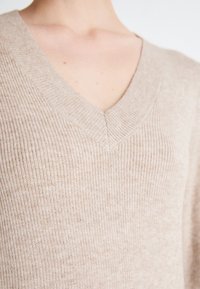 Pull à col en V beige clair avec texture côtelée, fabriqué en tissu doux et à la coupe décontractée, mettant en valeur l'encolure et les détails de couture.
