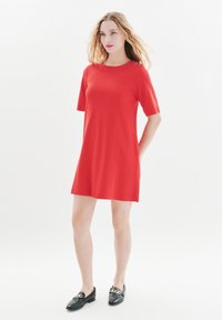 Rotes Kurzarmkleid mit lockerer Passform, mit dekorativen goldenen Knöpfen an der Schulter. Kombiniert mit schwarzen Slippern mit goldener Kettendetail.