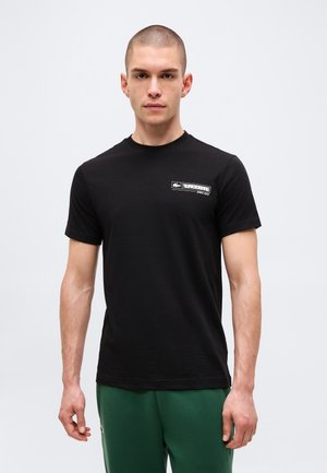 Giovane uomo che indossa una t-shirt nera Lacoste e pantaloni verdi, in piedi davanti a uno sfondo chiaro e semplice.