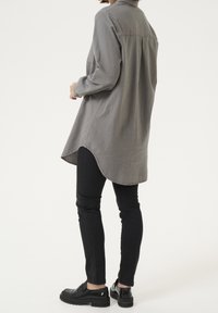 Camisa gris de manga larga con dobladillo curvado, cuello tipo button-down y una textura suave, combinada con jeans slim negros y zapatos negros.