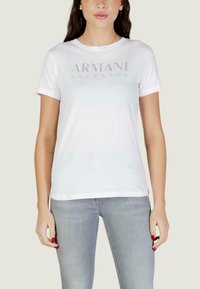 Femme portant un t-shirt blanc Armani Exchange et un jean gris clair, debout les bras détendus le long du corps devant un fond uni.