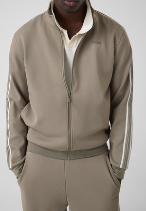 Mann trägt eine taupefarbene Trainingsjacke mit Reißverschluss und passende Hose mit weißen Seitenstreifen, Hände in den Taschen, cremefarbenes Shirt darunter, neutraler Hintergrund.
