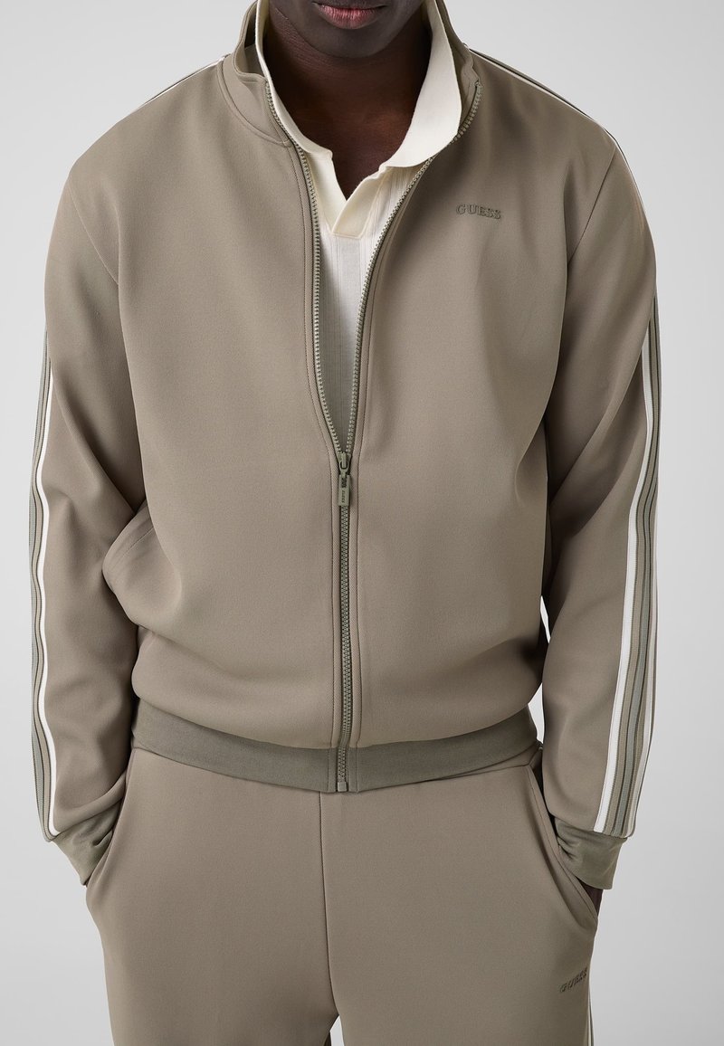 Homme portant une veste de survêtement taupe zippée et un pantalon assorti avec des bandes latérales blanches, mains dans les poches, chemise crème en dessous, fond neutre.