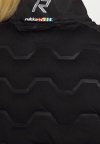 Svart jacka bak med präglat hexagonmönster, "R"-logotyp med vita prickar och en flerfärgad "rukka"-logotyp nära kragen.