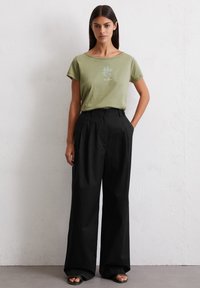 Olivengrünes Baumwoll-T-Shirt mit floraler Gestaltung, kombiniert mit hochgeschnittenen schwarzen weiten Hosen und offenen Sandalen.