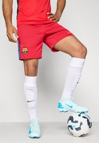 Punased jalgpallitossud siniste aktsentidega ja Barcelona embleemiga; koos valgete sokkide ja helesiniste spordijalatsitega, seistes mustrilisel jalgpallil.