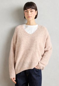 Helles rosa Oversized-Pullover mit V-Ausschnitt, aus weichem, flauschigem Material. Lange Ärmel und eine lockere Passform. Über einem weißen Oberteil getragen.