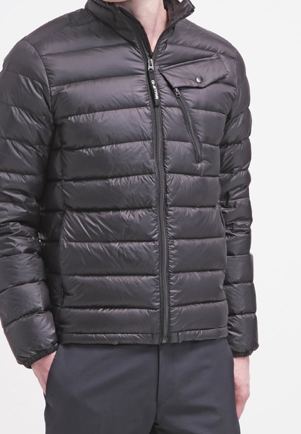 Chaqueta acolchada negra con diseño acolchado horizontal, cuello alto, cierre frontal con cremallera y un bolsillo en el pecho con cierre de presión. Material ligero.