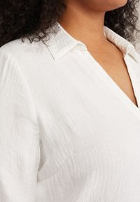 Camisa blanca de cuello en V, hecha de una tela texturizada, que presenta un ajuste relajado y material suave y ligero.