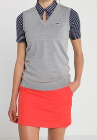 Pull sans manches gris à col en V porté sur un polo rayé bleu marine, associé à une mini-jupe corail vif. Logo Lacoste sur le pull.