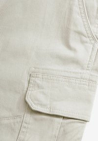 Gros plan sur un tissu beige avec une poche à rabat cousue, montrant des lignes de couture détaillées et un matériau en coton texturé.