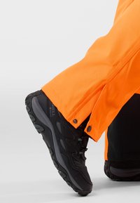 Zwarte gestructureerde sneaker met veters, gecombineerd met een oranje waterdichte broek met drukknoopsluitingen aan de zoom.