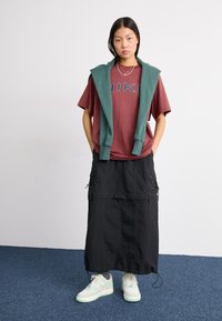 Camiseta roja con el logo de "NIKE", sudadera verde caída sobre los hombros, falda larga de cargo negra, zapatillas verde claro con acentos blancos.