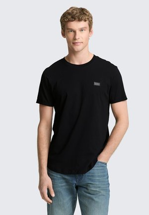 Junger Mann mit lockigem blondem Haar, der ein schlichtes schwarzes T-Shirt und blaue Jeans trägt, steht mit einer Hand in der Tasche vor hellem Hintergrund.