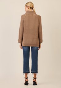 Maglione oversize a coste marrone con collo alto, maniche arrotolate e vestibilità rilassata, abbinato a jeans blu cropped e sandali neri con tacco.