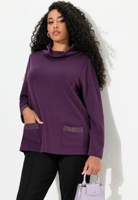 Top viola con collo a cowl e maniche lunghe, dotato di due tasche frontali con dettagli in strass, abbinato a pantaloni neri e una borsa lavanda.