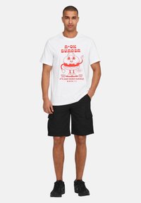 T-shirt bianca con grafica di un hamburger in stile cartoon rosso e testo, abbinata a pantaloni cargo neri e sneakers nere. Design semplice e casual.