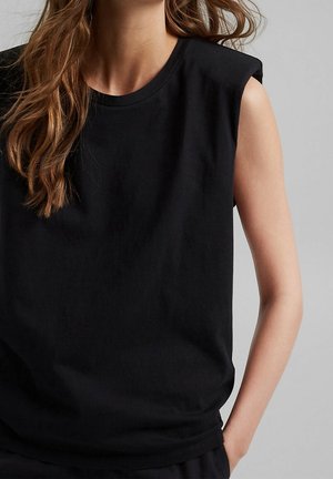 Vrouw met lang bruin haar die een mouwloos zwart topje draagt met gewatteerde schouders, één hand in de zak, tegen een grijze achtergrond.