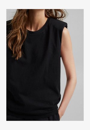 Kobieta z długimi brązowymi włosami, ubrana w czarny bezrękawnik z wyściełanymi ramionami, z jedną ręką w kieszeni, na szarym tle.