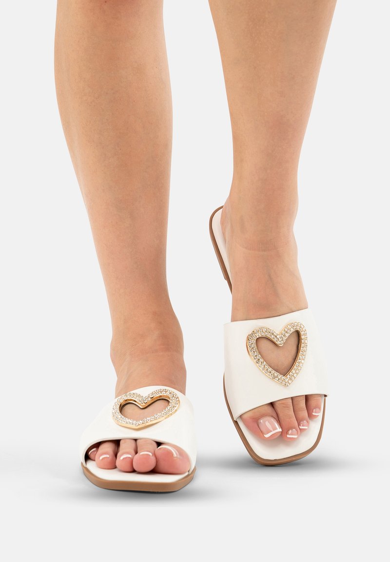 Sandali bianchi slip-on con una fascia larga caratterizzata da un cuore ritagliato in oro, decorato con strass. Texture liscia e suola piatta.