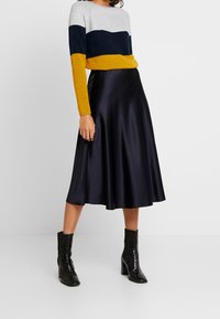 Maglione color block grigio, blu navy e senape, abbinato a una gonna nera in satin fluida e stivaletti neri con tacco. Le texture lisce sono ben evidenti.