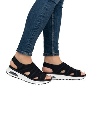 Black athletic sandals with open design, stretchy straps, and white sole featuring air cushioning. Worn with dark blue skinny jeans.