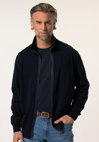 Tmavě modrý zip-up kardigan s vysokým límcem, přední kapsou a žebrovanými manžetami, který je nošen přes námořnickou košili a kombinován s modrými džíny.