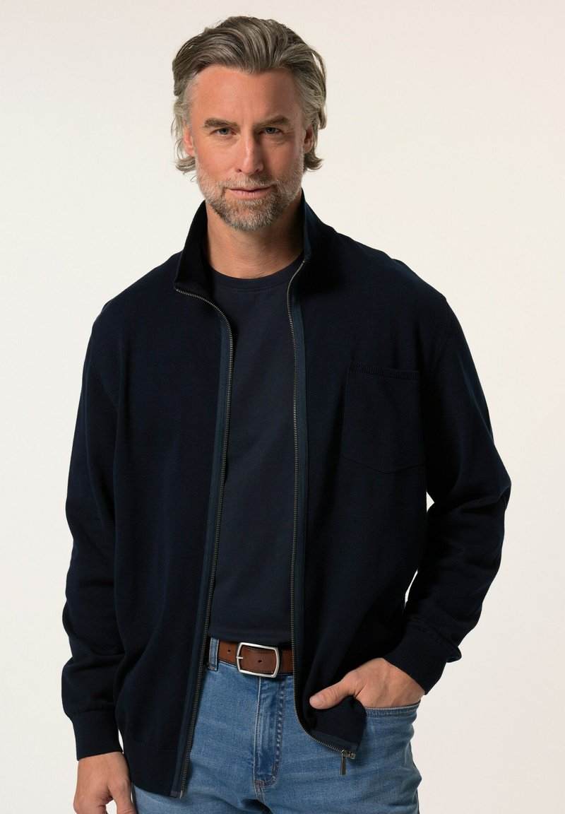 Tmavě modrý zip-up kardigan s vysokým límcem, přední kapsou a žebrovanými manžetami, který je nošen přes námořnickou košili a kombinován s modrými džíny.