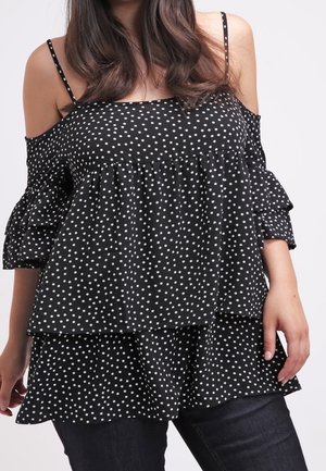 Femme portant un haut noir à épaules dénudées avec des pois blancs et des couches à volants, associée à un jean foncé sur un fond neutre.