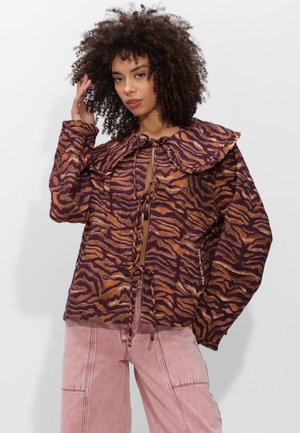 Vrouw met krullend haar die een bruin-zwart tijgergestreepte jas draagt met een grote kraag en vetersluitingen, gecombineerd met lichtroze broek.