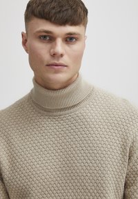 Beige turtleneck sweater with a textured pattern, ribbed collar, and knit fabric. Focus on design details, including stitchwork and shape.