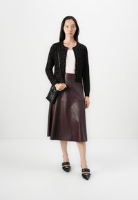 Cardigan noir à motifs avec des boutons ronds, top rose pâle, jupe midi en cuir bordeaux, et ballerines noires à bout pointu avec accents de boucle.