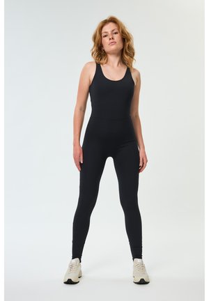 Žena s ramenatými červenými vlasy stojí, oblečená v černém přiléhavém overalu bez rukávů a béžových teniskách na bílém pozadí.