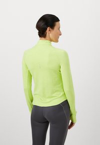 Vrouw met kort donker haar, gekleed in een felgroen sportshirt met lange mouwen en donkergrijze leggings, met de rug naar de camera gekeerd tegen een witte achtergrond.