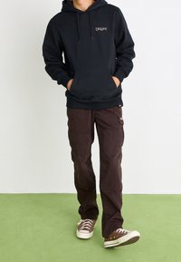 Sweat à capuche noir avec une poche avant et un logo, associé à un pantalon cargo marron avec plusieurs poches, et des baskets en toile marron.